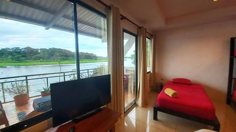 A cozy GuestHouse bedroom with a single bed in red bedding, TV, and large windows opens to a balcony overlooking Tortuguero's lush river scenery—perfect for relaxing after your adventures.