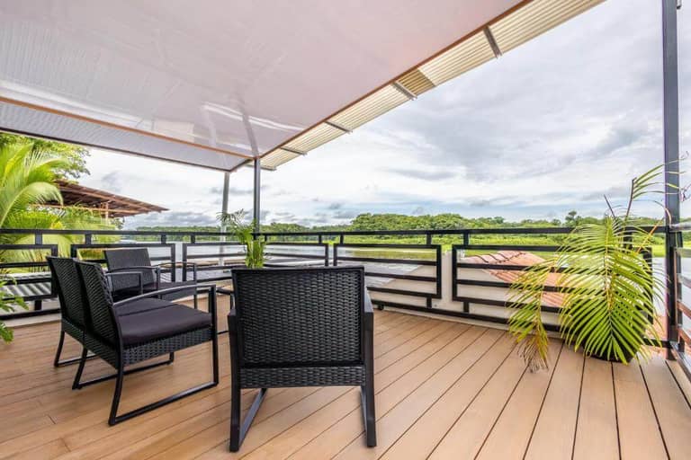 Outdoor terrace with wicker chairs, a table, potted plants, and a canopy roof, overlooking lush green trees and a cloudy sky.