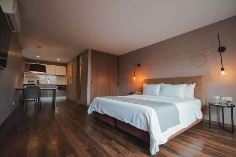 Modern Sites Hotel Medellin room with a large bed, side tables, wall lamps, wooden floor, and an open kitchenette in the background.