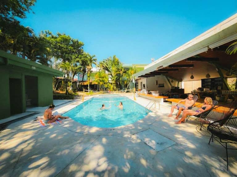 People relax around and swim in the outdoor pool at Selina La Fortuna, surrounded by palm trees and buildings on a sunny day.