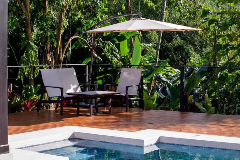 Two lounge chairs and a table sit under a large umbrella on a wooden deck beside the pool at Arenal Manoa Resort, surrounded by lush green tropical plants.