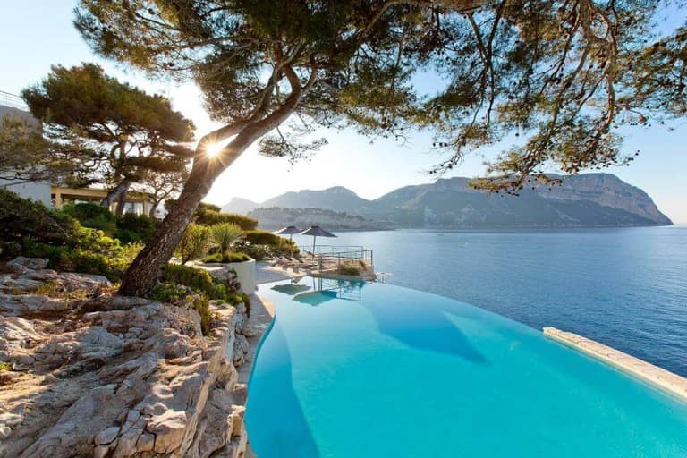 Infinity pool at Les Roches Blanches Cassis overlooking the ocean, with a rocky landscape, trees, and distant mountains under a clear sky at sunrise or sunset.
