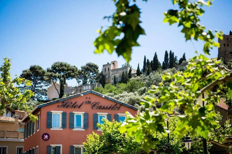 A pink hotel building with Hôtel Cassitel and The Originals Boutique branding is surrounded by green trees; a historic stone building sits atop a hill in the background under a clear blue sky.