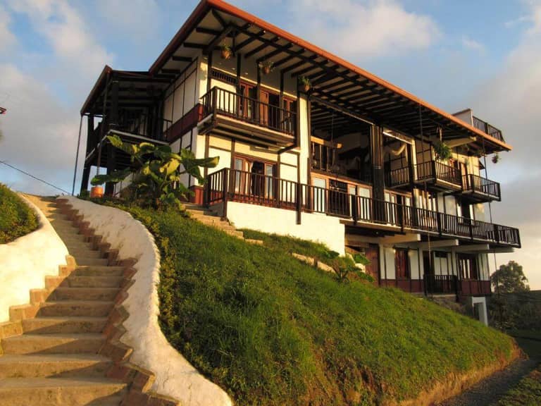 Two-story house with balconies and black railings sits on a grassy slope, with a curved concrete staircase leading up to the entrance of this inviting boutique hostel.