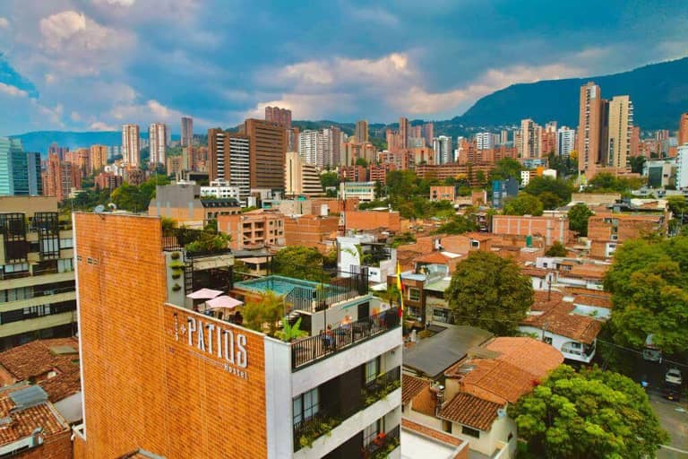 A cityscape view of Medellín, Colombia, with Los Patios Hostel Medellin nearby, showcasing brick buildings, high-rise apartments, green trees, and mountains in the background under a cloudy sky.