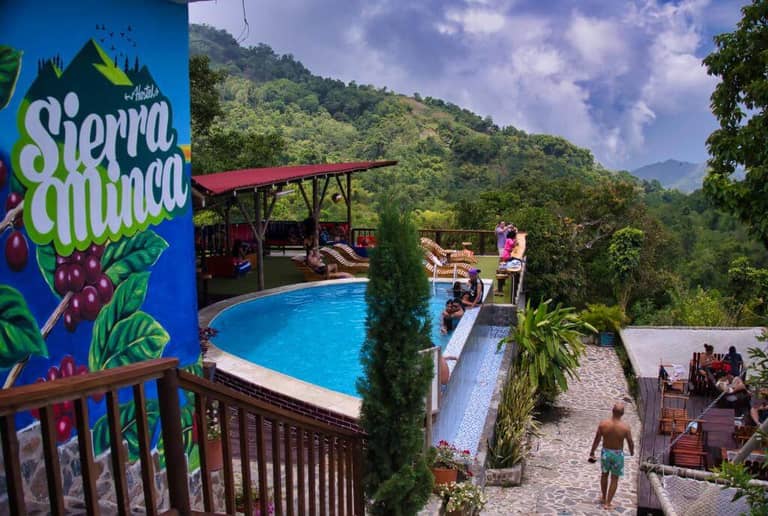 Outdoor pool area at Hostal Sierra Minca hillside resort with people relaxing; lush green mountains and a cloudy sky in the background; vibrant Sierra Minca mural on a blue wall.