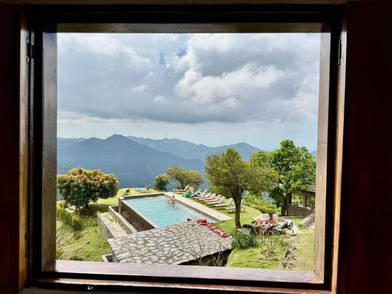 View through a window of the Mundo Nuevo Eco Lodge's outdoor pool, surrounded by lounge chairs, trees, and mountains under a cloudy sky—an ideal retreat for sustainable tourism enthusiasts.