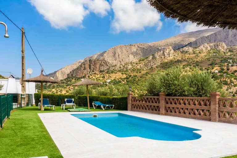 Outdoor swimming pool with sun loungers and straw parasols at Lagar del Chorro, set against a stunning backdrop of mountains and greenery under a partly cloudy sky.