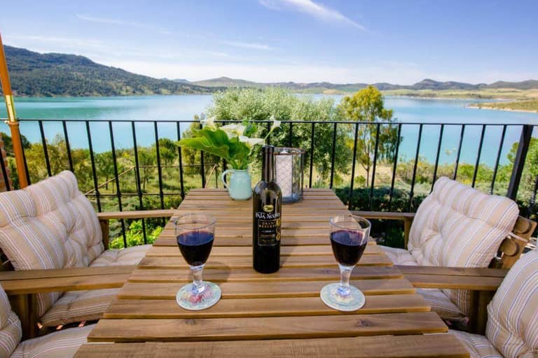 A wooden table with two glasses of red wine and a wine bottle sits on a balcony overlooking a lake and mountains under a clear sky.