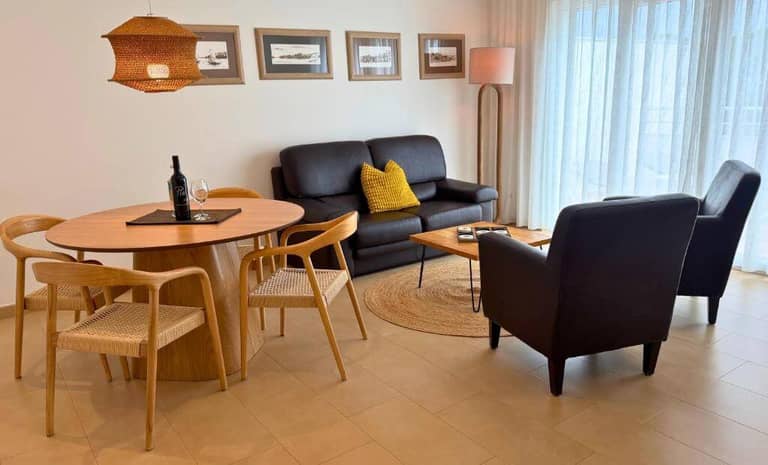 A modern living room at Pine Tree Apartments features a round wooden dining table with four chairs, a black sofa, armchair, floor lamp, framed art, and a large window with sheer curtains.
