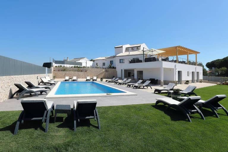 Modern white villa, Sunfield Guesthouse, features a large rectangular outdoor swimming pool surrounded by lounge chairs on a tiled deck and grass, all set under a clear blue sky.
