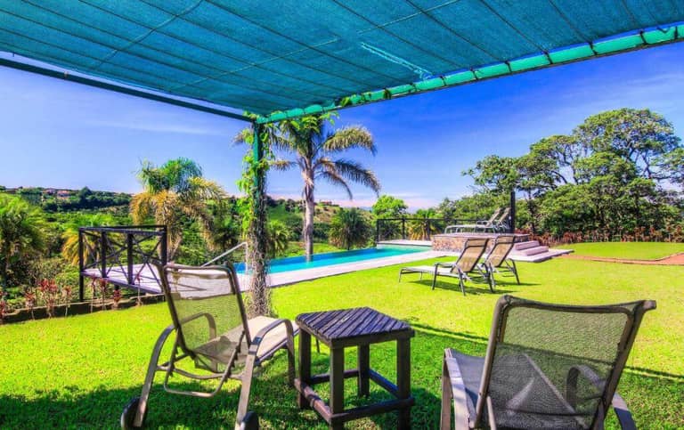 Two lounge chairs and a small table sit on a grassy lawn under a shade canopy at Hotel Mango Valley, overlooking a pool and palm trees with a scenic, sunny landscape in the background.