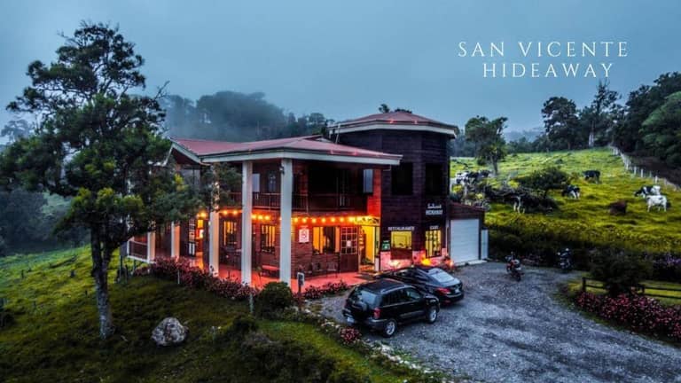 A two-story lodge with glowing lights sits in a green, misty rural landscape, with several cars parked outside. Text reads Hotel San Vicente Hideaway.