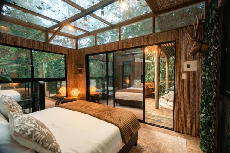A cozy BubbleSky bedroom with glass ceiling and walls, a bed with white linens, warm lighting, and a view of a wooden deck surrounded by greenery—perfect for glamping in Guatapé.