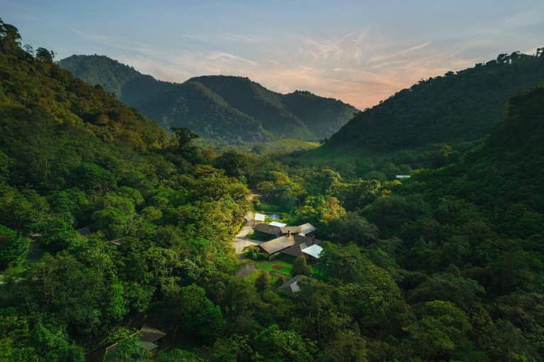 Aerial view of El Silencio Lodge, a serene spa and lodge nestled among dense green forest and hills under a partly cloudy sky at sunset.