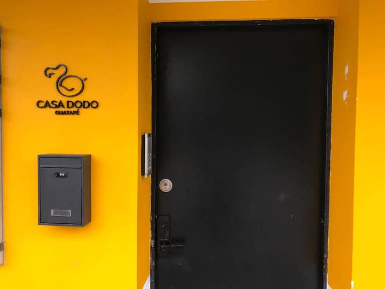A black door with a mailbox is set in a bright yellow wall in Guatape; a sign reads Casa Dodo Gamaté with a stylized dodo bird logo beside it.