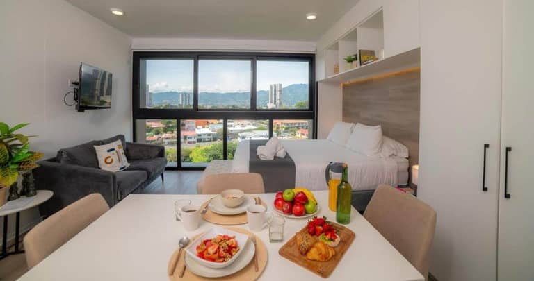 Modern studio apartment in San Jose with a double bed, small sofa, dining table set for breakfast, large window offering city views, and minimalist decor—perfect for a cozy stay at Train Suites.