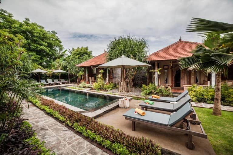 A tropical villa at Global Royal Pemuteran features a rectangular swimming pool, sun loungers with towels, umbrellas, lush greenery, and traditional red-roofed buildings under a cloudy sky.
