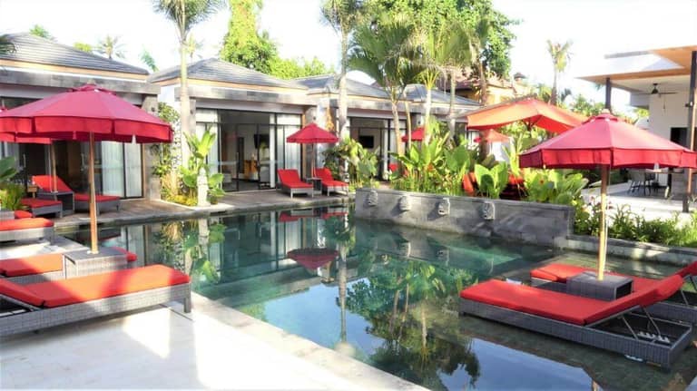 Modern outdoor pool area at Le Chien Rouge in Lovina, featuring red lounge chairs and umbrellas, lush tropical plants, and villa-style buildings in the background.