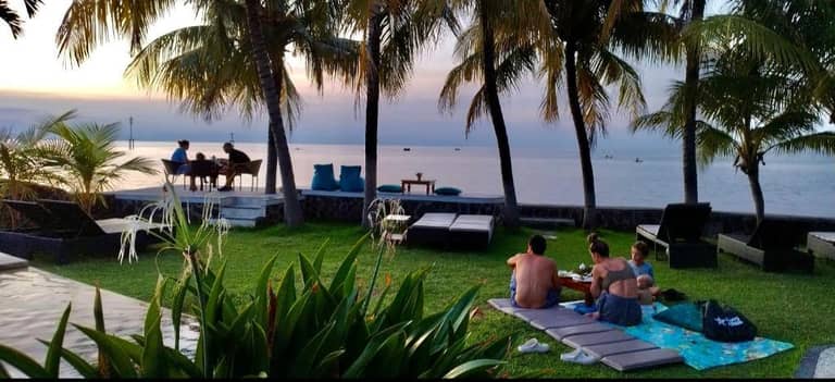 People sit and relax on lounge chairs and mats on a grassy area by the sea at 1000 Dream Bungalow Lovina, with palm trees swaying and a stunning sunset in the background.
