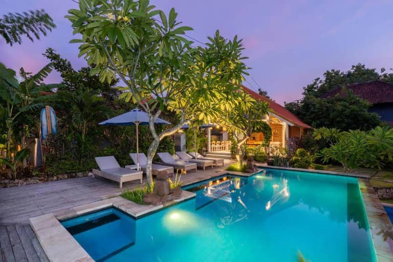 A tropical backyard at Le Biu Lembongan features a swimming pool, lounge chairs, an umbrella, lush greenery, and a small house glowing under the sunset.