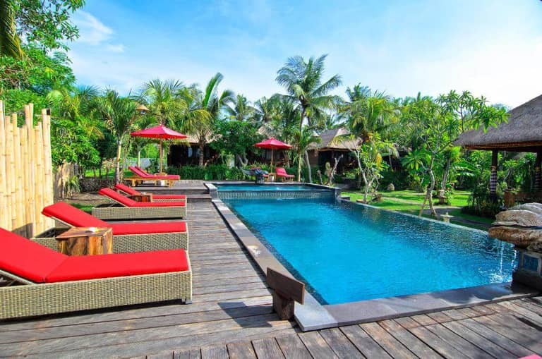 Outdoor swimming pool with clear blue water at Jingga Villas, surrounded by a wooden deck, red cushioned lounge chairs, green plants, and palm trees under the sunny Lembongan sky.