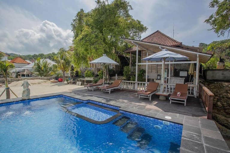 Outdoor pool with blue tiles, sun loungers, and umbrellas on a wooden deck beside sandy beach and charming Wooden Cottages under a partly cloudy sky.