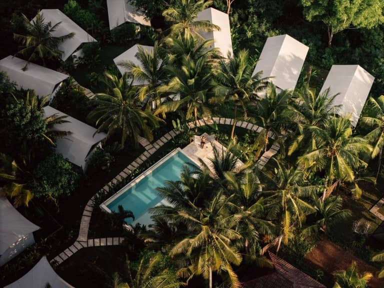 Aerial view of a rectangular swimming pool surrounded by palm trees and white-roofed huts in a tropical Glamping Penida setting.