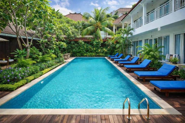 Rectangular outdoor swimming pool at Easy Surf Camp, Kuta, surrounded by blue lounge chairs, tropical plants, and a two-story building with balconies under a partly cloudy sky.