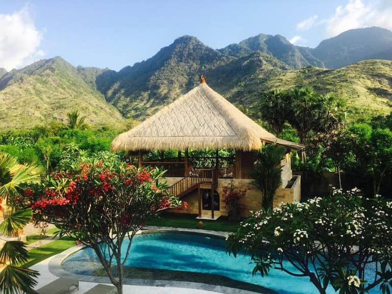 A thatched-roof pavilion at Sun Suko Pemuteran overlooks a curved swimming pool surrounded by tropical plants, with green mountains in the background under a clear sky.