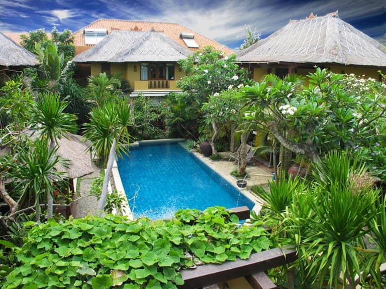 A rectangular swimming pool at D'Sriwing Villa Kuta is surrounded by tropical plants and traditional-style buildings under a partly cloudy sky, offering relaxing Kuta accommodation.