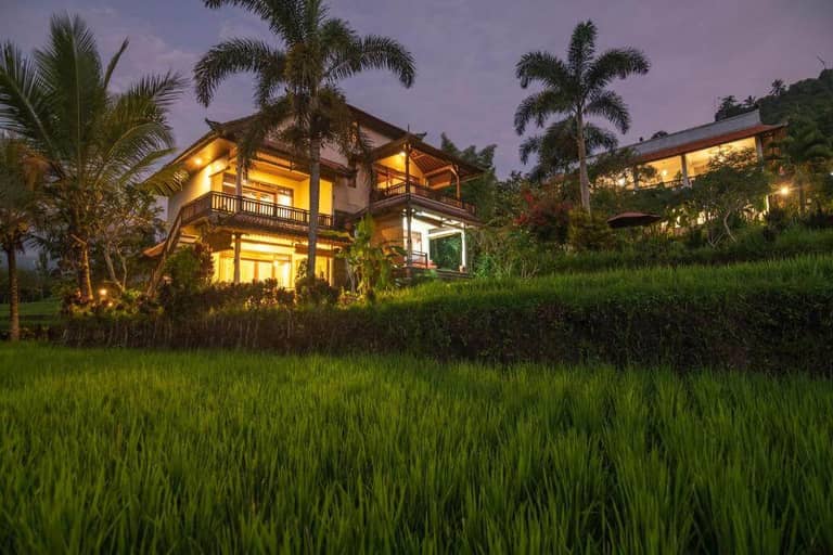 Two-story house with illuminated balconies at dusk, surrounded by palm trees and lush green rice fields near Lesong Hotel Munduk.