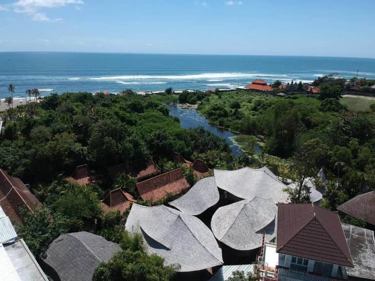 Aerial view of a coastal area with dense greenery, scattered buildings with sloped roofs, a small river, and the ocean in the background—an idyllic scene reminiscent of a Bali resort like ZIN Canggu Resort under a clear blue sky.