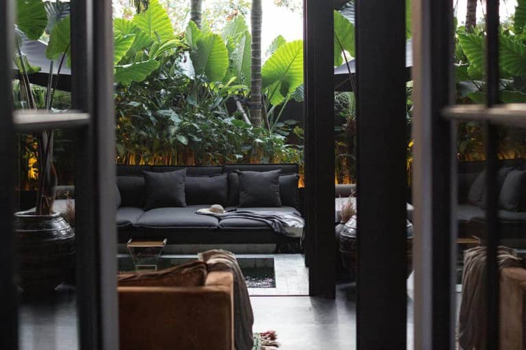 Outdoor patio with dark cushioned seating, a coffee table, and lush green plants in the background, viewed through open glass doors—evoking a relaxed Bohemian Canggu vibe.