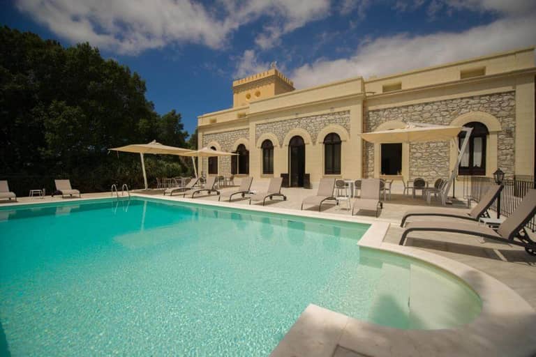 Outdoor swimming pool with lounge chairs and umbrellas in front of Villa le Torri Trapani's beige stone building under a partly cloudy sky.