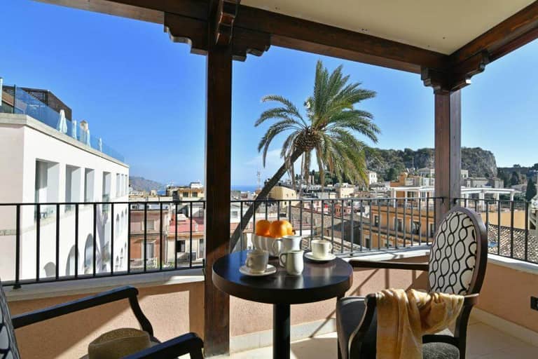 Covered balcony at Hotel Le Chevalier with a table set for coffee, two chairs, and a palm tree, overlooking Taormina’s cityscape with buildings and distant hills under a clear blue sky.