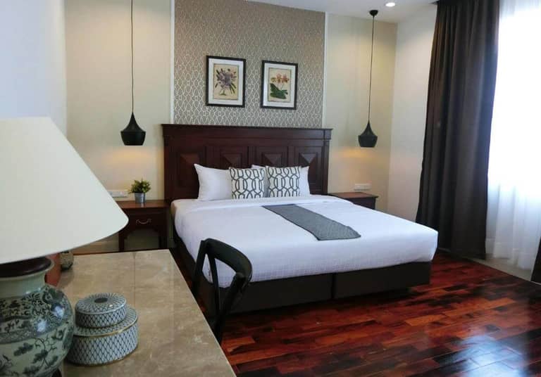 A neatly made double bed with patterned pillows and a gray runner, two bedside tables, pendant lights, a desk with a chair, and framed art on the wall in the bright Bloom Boutique Hotel bedroom.