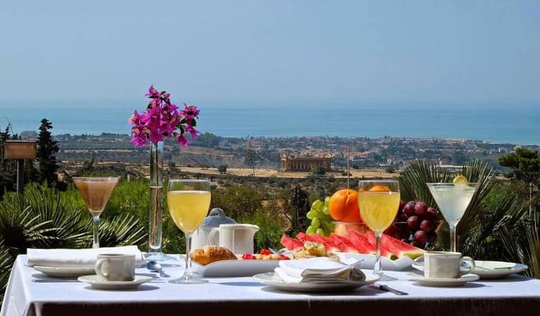 A breakfast table with juice, coffee, fruit, and pastries at Colleverde Park Hotel overlooks a scenic landscape with Agrigento’s coastal city and the sea in the background.