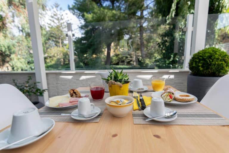 A breakfast table set for two with pastries, cold cuts, juice, and coffee on a patio at Apollo B&B in Agrigento, surrounded by lush greenery.