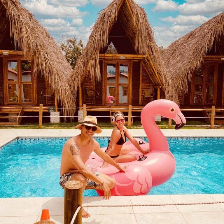 Two people in swimsuits enjoy a sunny day by the pool; one sits on a large pink flamingo float, with wooden huts and a stunning Palomino Sunrise sky in the background.