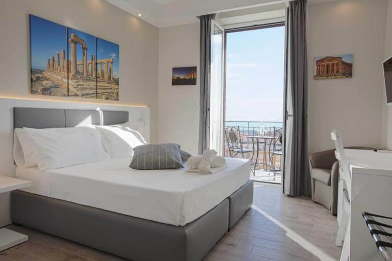 Modern Esseneto hotel room with a double bed, wall art of ancient ruins, desk, and an open balcony door revealing a sea view and outdoor seating in Agrigento.