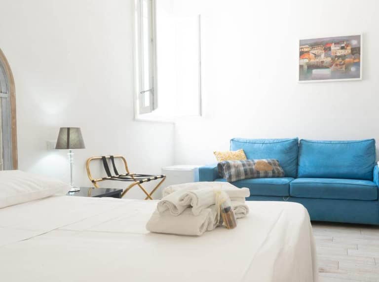 Bright Favignana bedroom with a neatly made bed, folded towels, blue sofa, open window, small lamp, luggage rack, and a framed picture on the wall create a welcoming Poggiamico ambiance.