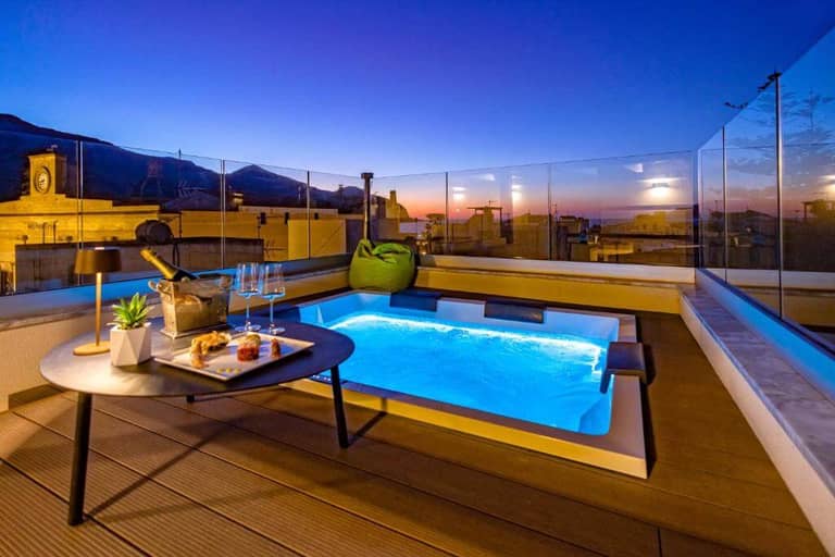 A rooftop terrace at dusk at Mazzini Favignana features a lit hot tub, a table with drinks and snacks, a green bean bag chair, and cityscape views enclosed by glass panels.