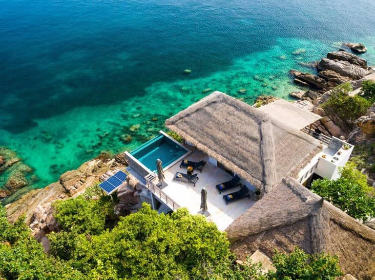 Aerial view of a thatched-roof villa at Cape Shark Villas with an infinity pool overlooking Koh Tao’s clear turquoise ocean, surrounded by rocks and lush greenery.