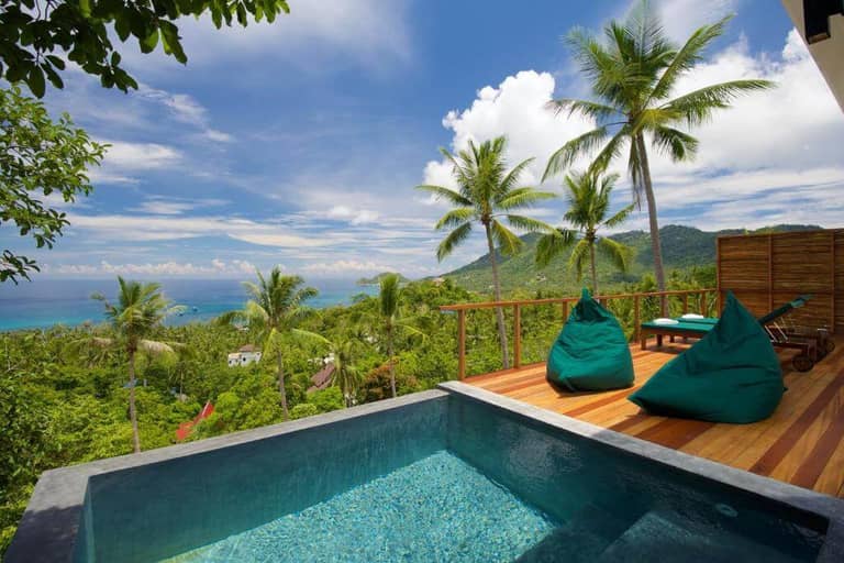 Infinity-edge pool with two green bean bag chairs on a wooden deck overlooks lush tropical trees, hills, and the ocean under a blue sky at Overthemoon Koh Tao—a stunning Koh Tao hotel retreat.