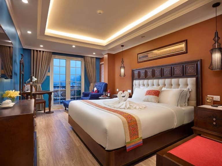 A neatly made Sapa Eden Boutique Hotel room with a large bed, swan towel art, wooden furniture, a blue armchair, a desk, and large windows with curtains revealing a scenic view outside.
