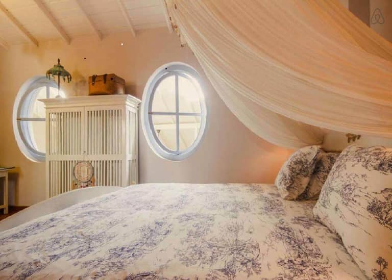 A cozy bedroom with patterned bedding, round windows, a white wardrobe, wicker baskets, and a draped canopy evokes the charm of Galle’s Jardin du Fort.