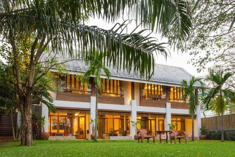Two-story modern guesthouse with large windows, balcony, and patio, surrounded by palm trees and a green lawn, viewed from the garden—a serene retreat reminiscent of Baan Tye Guesthouse in Ayutthaya.