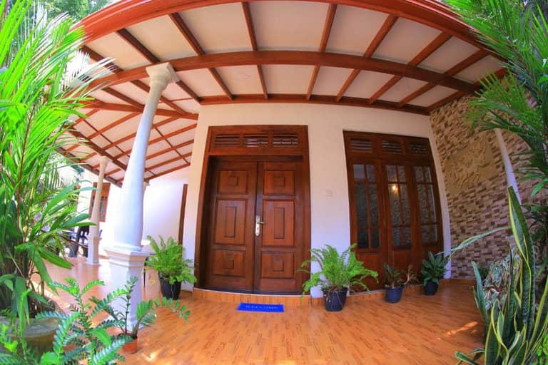 A wooden double door entrance at Abinus Place, Dikwella, with potted plants on a tiled porch, white columns, and a partial stone wall under a wooden ceiling.