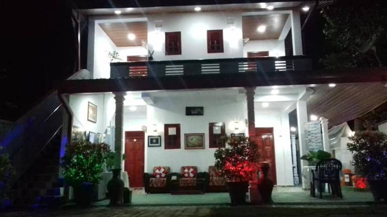 Two-story house at night with a covered porch, red and green lights illuminating potted plants, and outdoor seating visible on the right side—an inviting scene reminiscent of Green Village Dambulla, a tranquil Dambulla hotel.